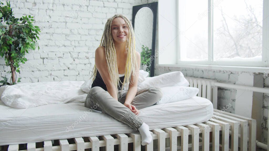 A girl with light dreadlocks sits on a bed with white sheets. She smiles and straightens her hair