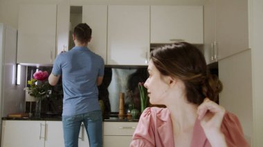 Genç bir bayan evde çalışıyor ve kocası da arka planda ev işlerini yapıyor. 