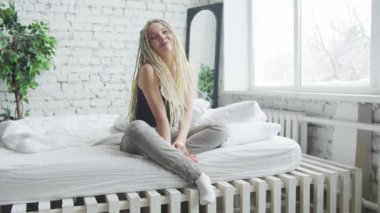 Hafif rastalı bir kız beyaz çarşaflı bir yatakta oturuyor. Gülümser ve saçlarını düzeltir.