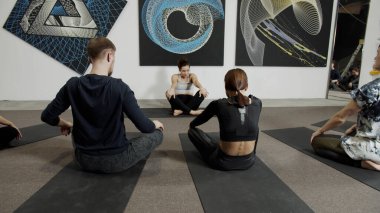 Genç bir kadın bir takımla yoga yapıyor, spor kıyafetleri giyiyor, siyah minderlerin üzerinde oturuyor.
