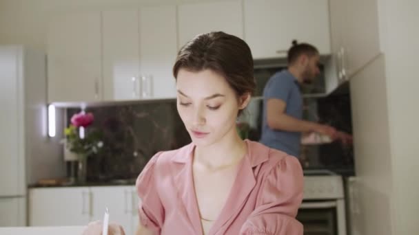 Eine junge Frau arbeitet zu Hause und ihr Mann erledigt im Hintergrund die Hausarbeit — Stockvideo
