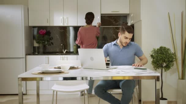 Un hombre serio está trabajando en casa y su esposa lo está mirando y bebiendo una taza de agua — Vídeos de Stock