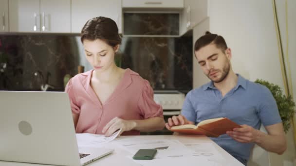 A young woman is working and home and her husband is reading a book — стоковое видео