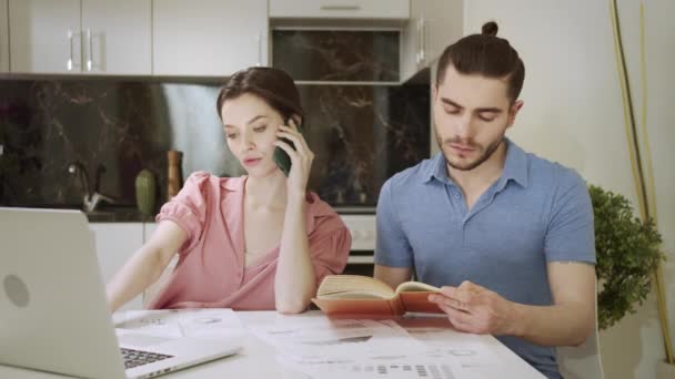 Egy fiatal nő beszél telefonon munka közben, és a férje könyvet olvas. — Stock videók