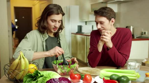 Um homem atraente está misturando a salada e conversando com seu amigo — Vídeo de Stock