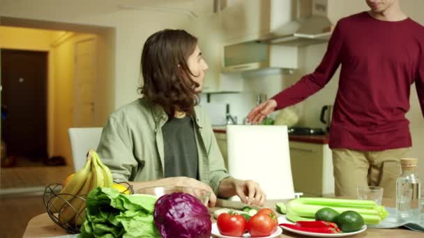 En ung man går in i ett kök medan hans vän lagar sallad. — Stockvideo