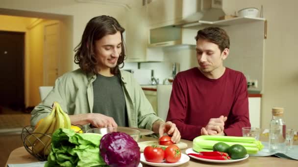 En ung man skär tomater för en sallad och pratar med sin vän. — Stockvideo