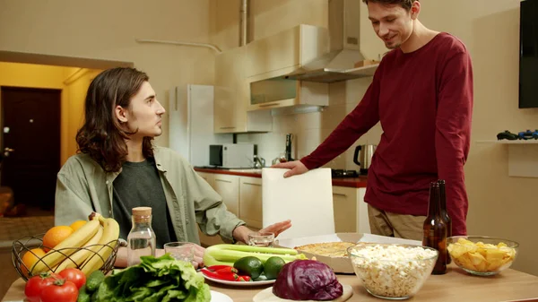 Uno de los amigos está sentado en la mesa mientras su amigo entra en la cocina. — Foto de Stock