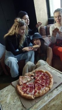 Farklı saç stilleri olan sportif gençler kanepede oturmuş pizza yiyorlar.