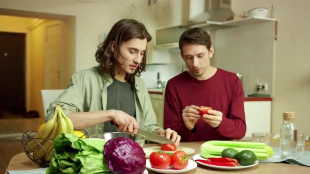 Un jeune homme coupe des tomates pour une salade et parle à son ami — Video