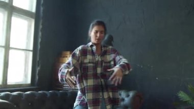 Genç kız dans ediyor. Stüdyoda karanlık duvarlar ve kanepe var.