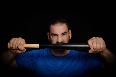 Küstah pozu veren mavi tişörtlü sakallı adam siyah bir arka planda beyzbol sopasıyla poz veriyor. Tehdit konsepti.