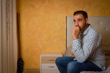 Çekici sakallı adam evdeki yatak odasında üzgün ve üzgün. 30-40 yaş arası dramatik bir hayat tarzı portresi, yatakta oturan yakışıklı bir adam, endişeli ve çaresiz. Depresyon kavramı