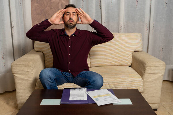 Беспокойный и отчаявшийся молодой человек, рассчитывающий домашние расходы, сидя дома на диване. Концепция финансовых проблем