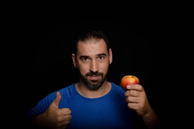 Mavi tişört giymiş sakallı bir adam ve elinde siyah arka planda duran bir elma var. Sağlıklı diyet kavramı