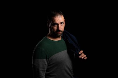 Omzunun üstünden sarkan mavi ceketli çekici sakallı adam siyah arka planda yalnız poz veriyor. Moda konsepti