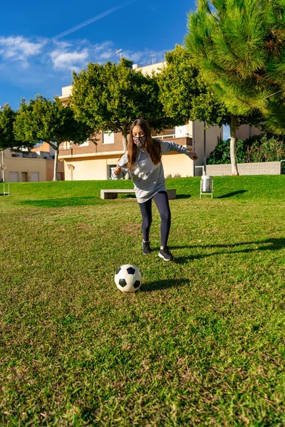 芝生の上でサッカーをしている美しい少女は 天然芝の美しい公園でスポーツをしている間 コロナウイルスから身を守るためにマスクを着用しています 健康的なライフコンセプト — ストック写真