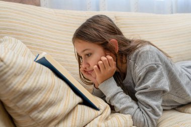 Oturma odasındaki kanepede kitap okuyan sevimli küçük kız. Seçici odaklanma. Öğrenme kavramı.