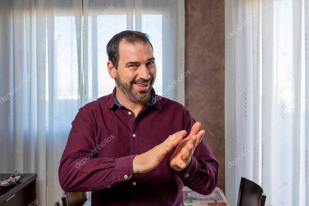 Young handsome man with a beard wearing a purple shirt clapping cheerfully, smiling and proud. Hands together. Party concept