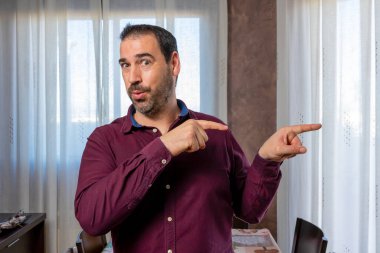 Mor gömlekli İspanyol yakışıklı adam beyaz perdelerle arka plandaki oturma odasında poz veriyor ve el kol hareketi yapıyor. Hareketli kavram