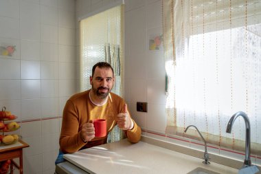 Turuncu kazaklı ve kot pantolonlu sakallı bir adam bir elinde kahve, diğer elinde de onaylı olarak parmağını kaldırıyor. İyimserlik kavramı