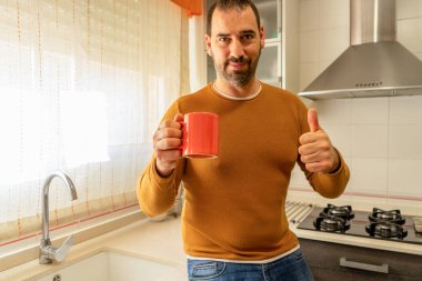 Turuncu kazaklı ve kot pantolonlu sakallı bir adam bir elinde kahve, diğer elinde de onaylı olarak parmağını kaldırıyor. İyimserlik kavramı