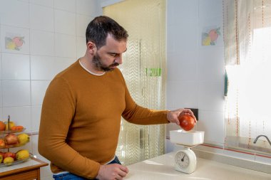 Turuncu kazaklı ve kot pantolonlu sakallı adam mutfak terazisine büyük bir kırmızı domates koyuyor. Sağlıklı gıda konsepti