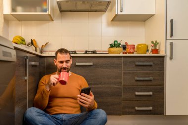 Turuncu kazaklı sakallı bir adam mutfakta oturmuş elinde bir fincan kahve tutarken cep telefonuna bakıyor. Ev kavramı