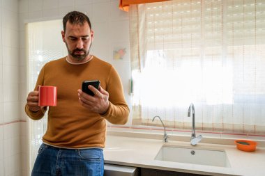 Turuncu kazaklı yakışıklı sakallı adam mutfakta kahve içerken akıllı telefona bakıyor. Teknoloji konsepti