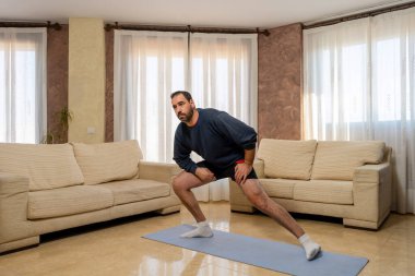 Koyu renk spor giyimli sakallı adam evdeki oturma odasında esneme hareketleri yapıyor. Egzersiz konsepti