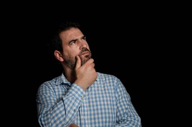 Portrait of handsome bearded man in a blue and white plaid shirt with his hand on his chin with thoughtful gesture on black studio background. Doubt concept