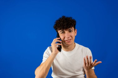 Mavi stüdyo arka planında izole edilmiş akıllı telefondan konuşan kıvırcık saçlı yakışıklı genç adam. Teknoloji konsepti