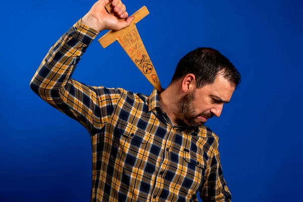 身穿黄色格子花衬衫的男人用一把木制剑划伤了他的背 这把剑被隔离在蓝色工作室的背景下 有趣的概念 — 图库照片