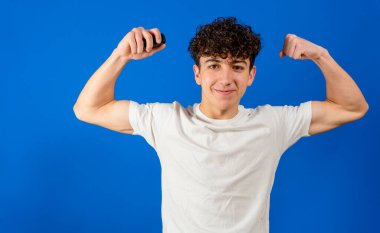 Çekici genç adam kameraya gülümserken kaslarını çekiyor. Mavi stüdyo arka planında izole edilmiş. Spor konsepti
