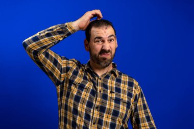 Bearded man in a yellow plaid shirt scratching his head with doubt gesture isolated on blue studio background. Riddle concept