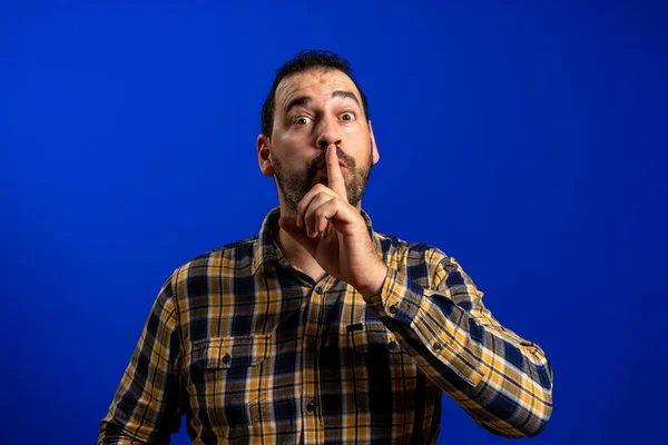 身穿黄色格子花衬衫 留着胡子的男人要求安静 把食指放在嘴唇上 隔离在蓝色的工作室背景下 Gesture概念 — 图库照片