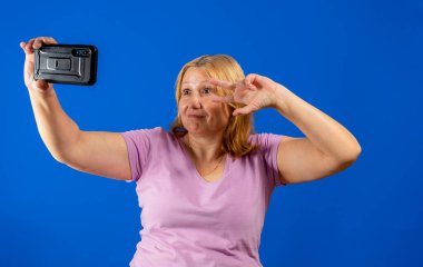 Mor tişörtlü orta yaşlı güzel bir kadın mavi stüdyo arka planında izole edilmiş akıllı telefonuyla selfie çekiyor. Teknoloji konsepti