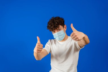 Kendini koronavirüsten korumak için ameliyat maskesi takan kıvırcık saçlı çekici bir adam ve mavi stüdyo arka planında parmakları var. Salgın konsepti