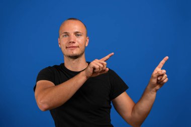 Çok kısa saçlı ve mavi gözlü çekici genç adam işaret parmaklarını mavi stüdyo arka planında izole edilmiş tarafa işaret ediyor. Hareket kavramı