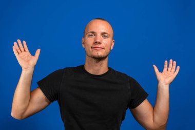 İzole edilmiş mavi arka planı olan siyah tişörtlü genç bir adam bir ilan vermek için avucunda hayali bir şey tutuyor. Hareket kavramı.