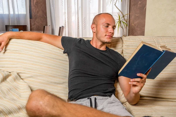 Jeune homme lisant un livre dans son salon assis sur le canapé. Concept d'étude. — Photo