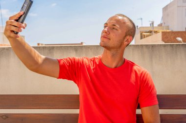 Kırmızı tişört giyen, ahşap bankta tek başına oturan ve cep telefonunda selfie çeken şık bir gencin açık hava portresi. İnsanlar ve modern teknoloji kavramı