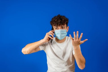Young man with curly hair and wearing a surgical mask to protect himself from coronavirus talking pissed off on his smartphone, isolated on blue studio background. New normal concept.