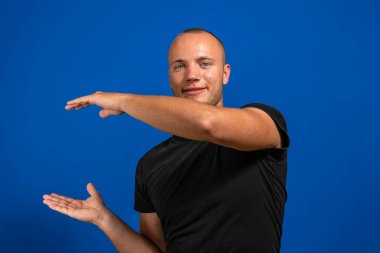 İzole edilmiş mavi arka planı olan siyah tişörtlü genç bir adam bir ilan vermek için avucunda hayali bir şey tutuyor. Hayal gücü konsepti