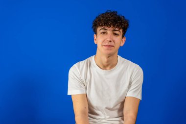 Kıvırcık saçlı, gülümseyen beyaz bir genç adam, iyi bir hayat yaşamaktan mutlu. Mavi stüdyo arka planında izole edilmiş. Memnuniyet kavramı
