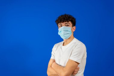 Kıvırcık saçlı ve cerrahi maske takan genç bir adam kendini koronavirüsten korumak için mavi stüdyo arka planında izole edilmiş. Yeni normal kavram.