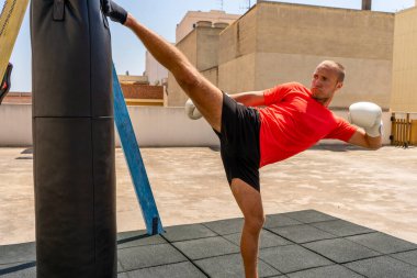 Siyah ve kırmızı spor giyimli beyaz bir adam kum torbasını tekmeliyor, en güçlülerle yarışmak için antrenman yapıyor. Eğlence konsepti