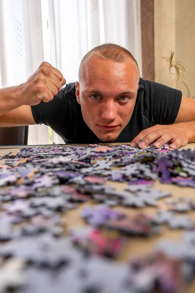 Attractive teenage caucasian man in a black t-shirt assembling a puzzle in the living room at home. Training concept — Stock Photo, Image