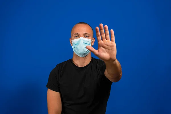 Alarming scared panicking man in hygienic mask gesturing stop, afraid of coronavirus infection, respiratory illnesses such as flu, 2019-nCoV, Covid-19, ebola. indoor shot isolated on blue background.