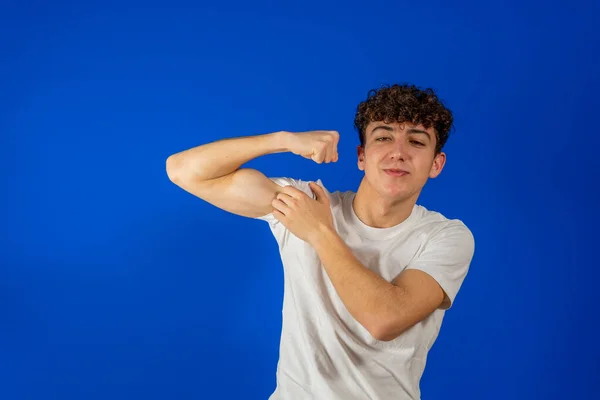 Trots man wijst naar zijn biceps en kijkt naar de camera. Portret van knappe hipster krullende jongeman in klassiek casual wit shirt. Indoor studio schot geïsoleerd op blauwe achtergrond. — Stockfoto